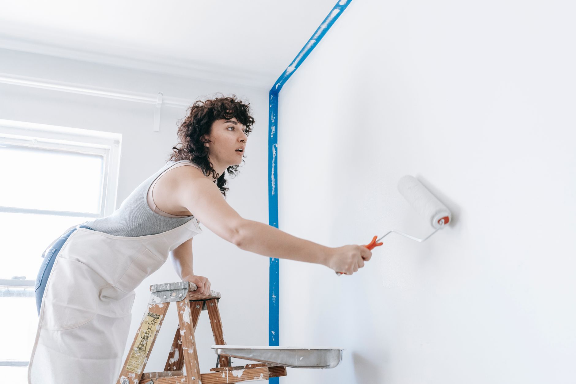 A woman stands on a ladder painting a wall white, part of a DIY home renovation