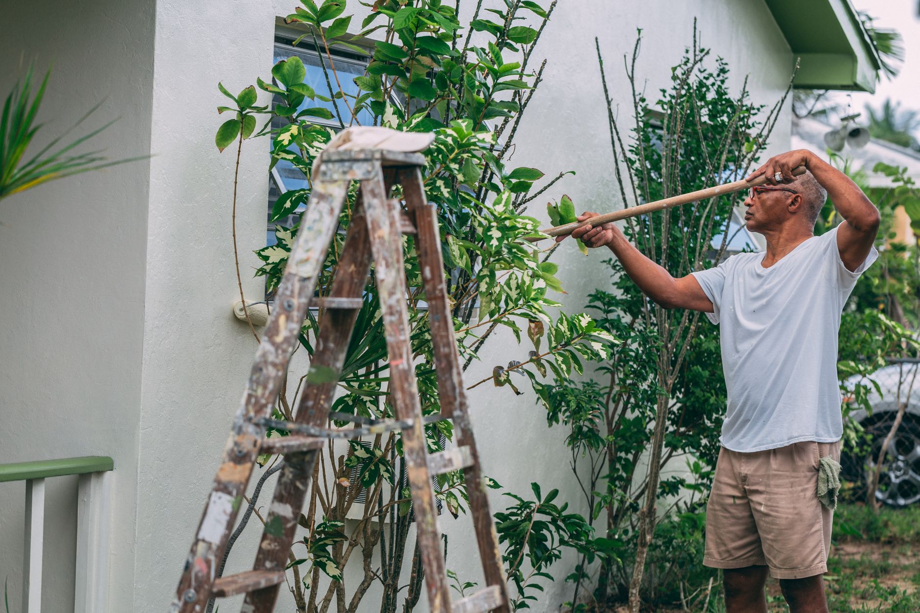 An adult man paints a house wall outdoors with a roller and ladder in a garden, showing the labor involved in an exterior repaint